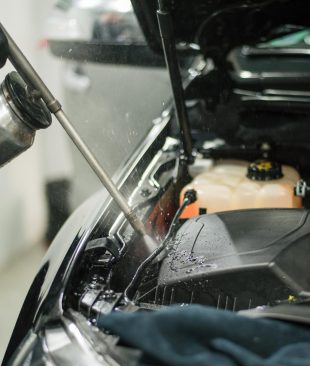 A worker wearing protective gloves is spraying a car engine with a can of cleaning solution